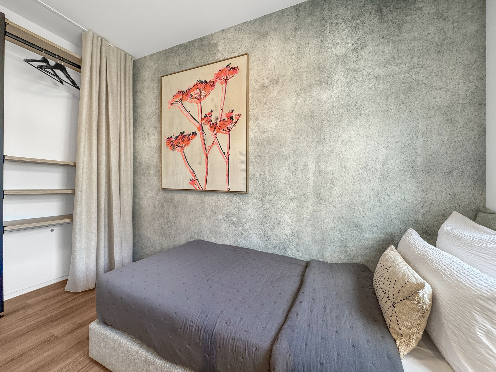 Boxspringbett vor einer Wand mit Sichtbeton-Optik, individueller Wandkunst und offenem Kleiderschrank
